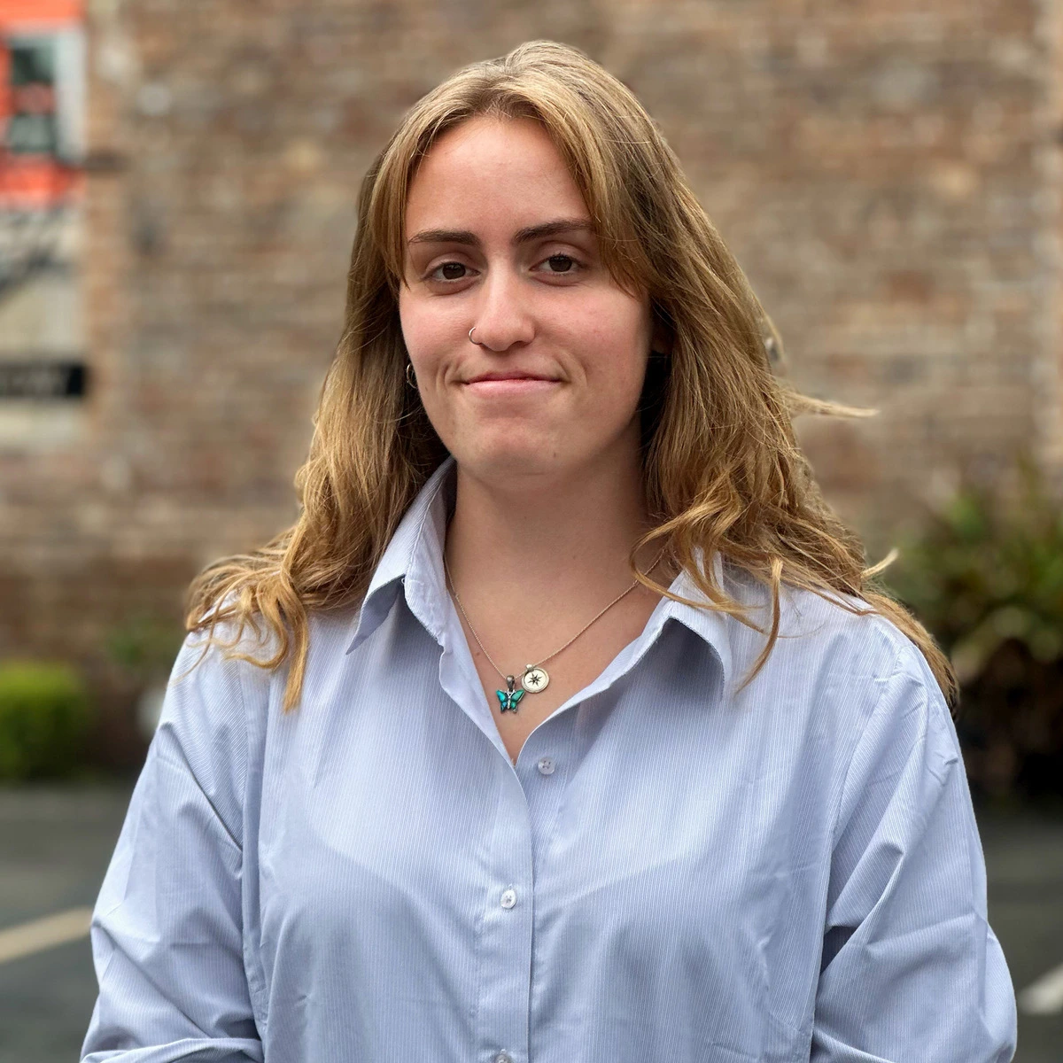 A woman with light brown hair wearing a light blue button-up shirt and a necklace stands outdoors, smiling slightly with a blurred brick wall and greenery in the background.