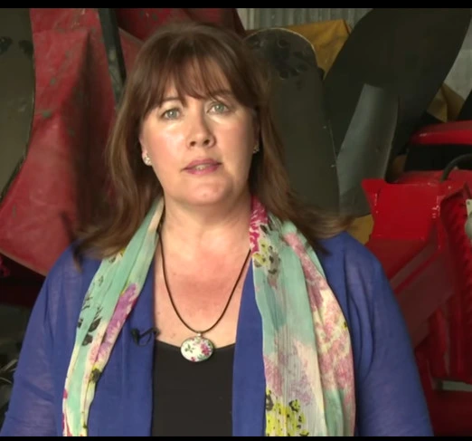A woman with brown hair, wearing a blue top and a floral scarf, stands indoors with colorful equipment or machinery in the background.